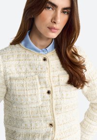 Giacca bianca con texture che presenta fili dorati, bottoni neri rotondi e una camicia blu sotto, caratterizzata da un motivo strutturato e tessuto.