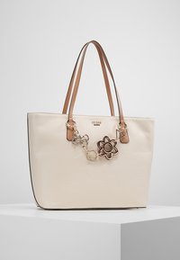 Kremowa torba tote z brązowymi skórzanymi uchwytami, wykonana z teksturowanego materiału. Posiada ozdobny charms z kwiatowym wzorem i logo.