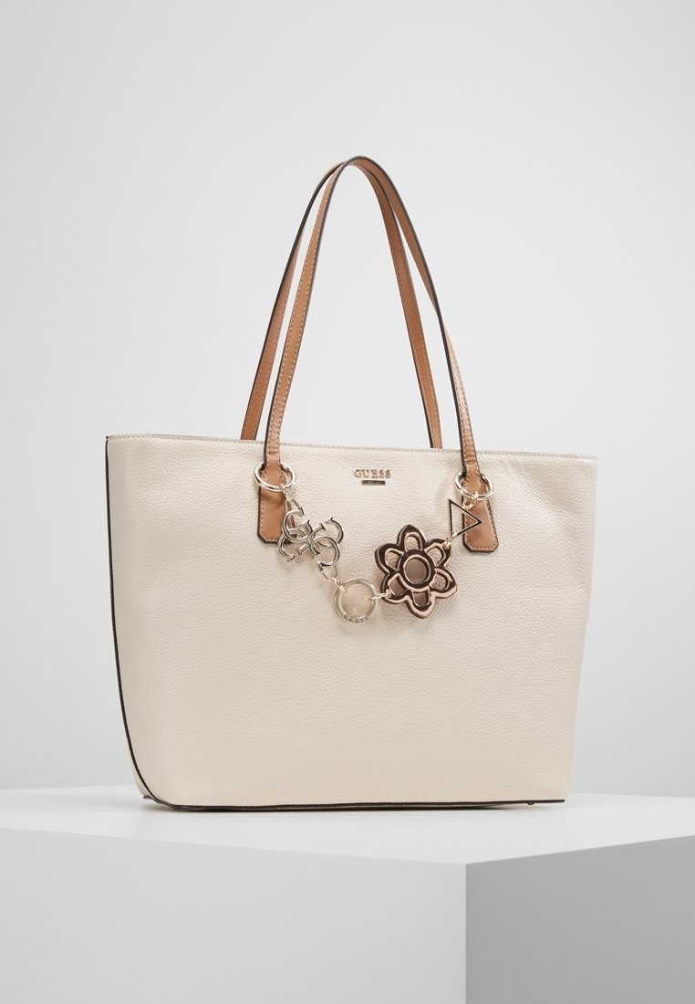 Kremowa torba tote z brązowymi skórzanymi uchwytami, wykonana z teksturowanego materiału. Posiada ozdobny charms z kwiatowym wzorem i logo.