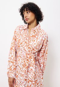 Robe blanche avec un motif abstrait orange, manches longues, col et ceinture à la taille pour un ajustement réglable.