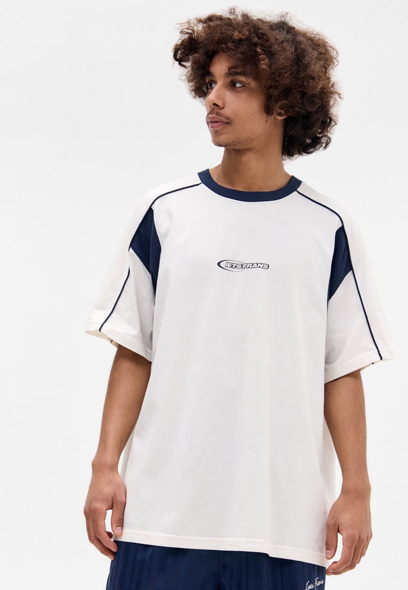 Witte oversized t-shirt met marineblauwe accenten en stiksels. Heeft een ronde hals en een logo op de voorkant. G gedragen met marineblauwe shorts.