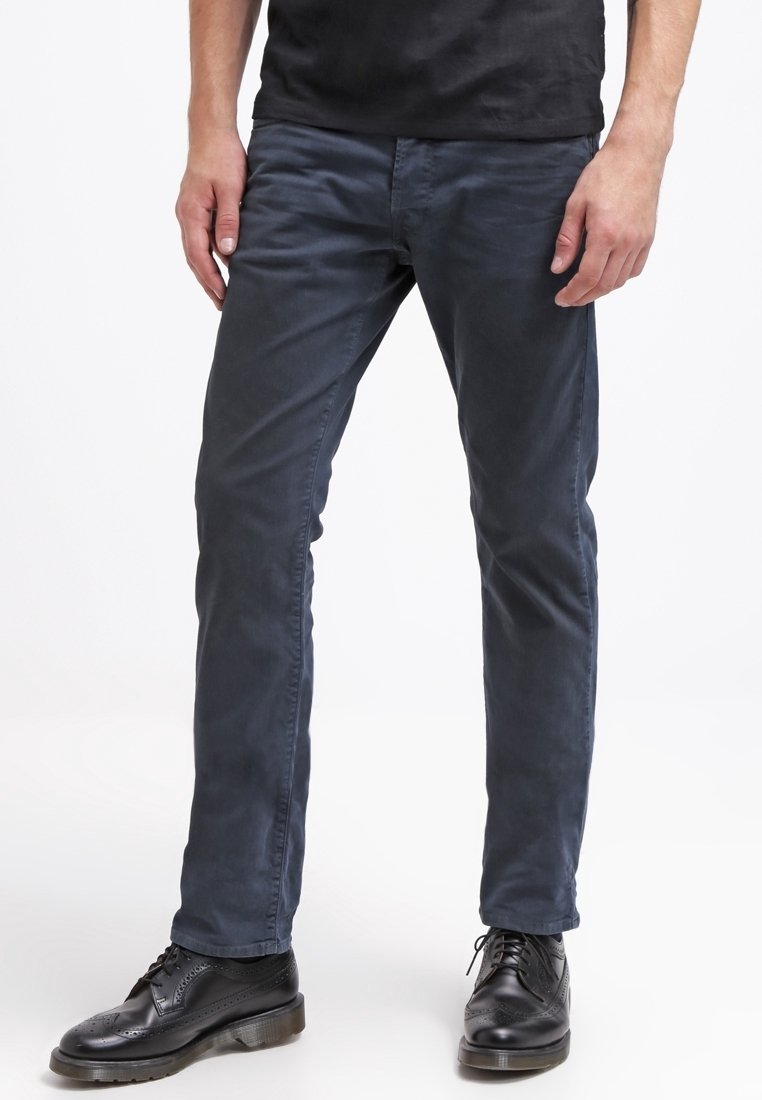 Homme portant un pantalon slim bleu marine foncé et des chaussures de ville en cuir noir, se tenant devant un fond blanc uni.