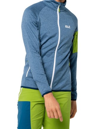 Uomo che indossa una giacca azzurra con zip e pantaloni verde lime e blu. La giacca presenta una tasca con zip sul braccio superiore e un logo bianco sul petto.