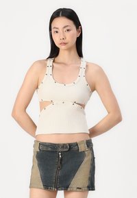 Top senza maniche beige a coste con design a intaglio e borchie metalliche, abbinato a una minigonna di jeans con pannelli a contrasto e zip frontale.