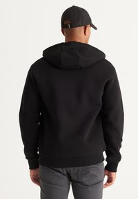 Zwarte hoodie met een trekkoord in de capuchon, ribgebreide cuffs en zoom, gemaakt van zacht materiaal. Draagt grijze broek en een donkere pet. Achteraanzicht.