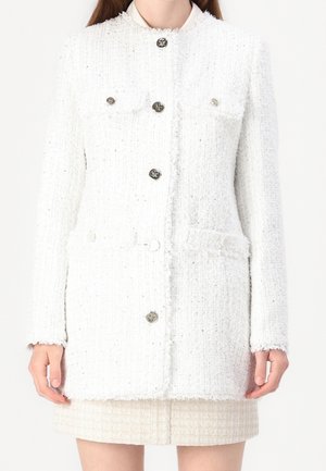 Femme portant une veste blanche texturée à manches longues boutonnée avec des poches à rabat sur une jupe courte assortie.