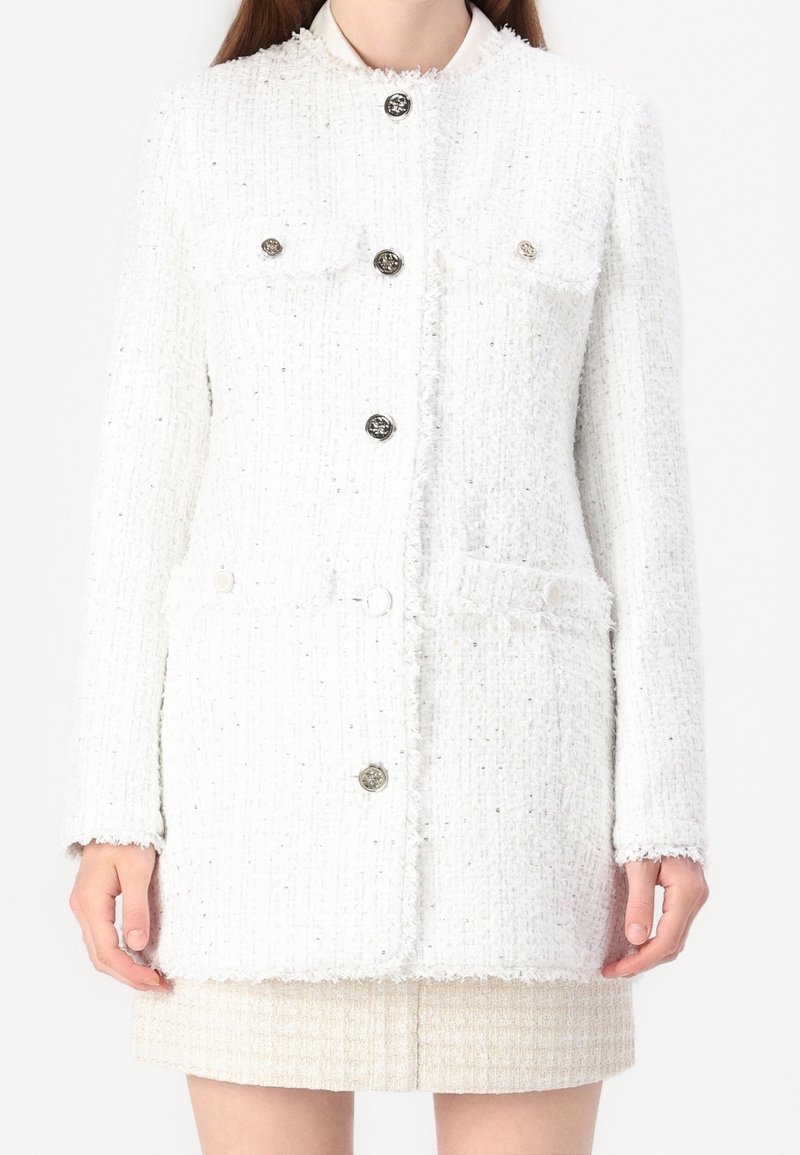 Femme portant une veste blanche texturée à manches longues boutonnée avec des poches à rabat sur une jupe courte assortie.