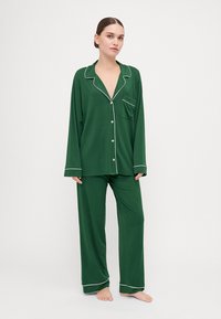 Green pajama set made from soft fabric. Features a button-up top with a pocket and contrasting white piped edges. Loose-fitting pants.