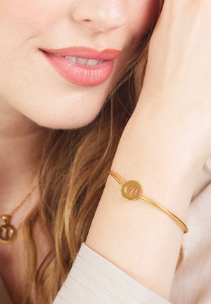 Bracelet doré avec un charme circulaire à découpe, finition métallique lisse, porté au poignet, sur un fond neutre.