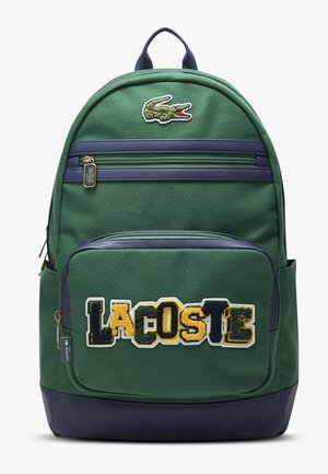 Zielono-granatowy plecak z przednią kieszenią na suwak, na której znajduje się wielokolorowy napis "LACOSTE" i logo krokodyla nad górnym suwakiem.