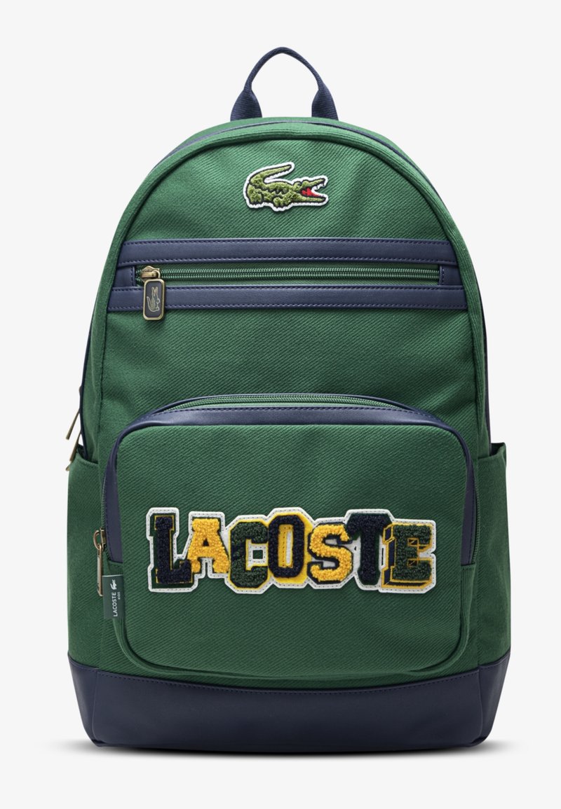 Sac à dos vert et bleu marine avec poche avant zippée affichant les lettres multicolores "LACOSTE" et le logo crocodile au-dessus de la fermeture éclair supérieure.