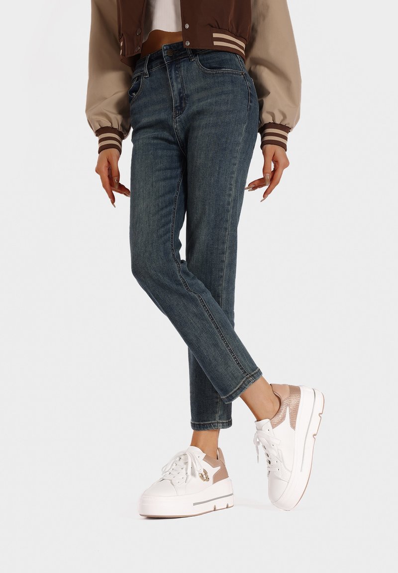 Sneakers bianchi con un accento taupe testurizzato, jeans in denim e una giacca bomber marrone e beige con polsini a righe. Mostra di un outfit casual.