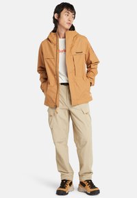Brun vattentät jacka med huva, två framfickor och Timberland-logotyp, tillsammans med beiga cargobyxor och svarta-orange vandringsskor.