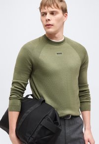 Jeune homme portant un pull BOSS vert olive, pantalon gris, tenant un sac de voyage noir, regardant sur le côté devant un fond uni.