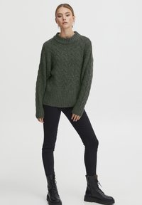 Dunkelgrüner Strickpullover mit Rippenbund und hohem Kragen, getragen über schwarzen Slim-Fit-Jeans und schwarzen Schnür-Ankle-Boots.