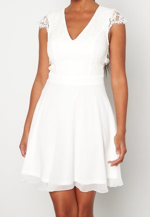 Femme portant une robe blanche sans manches avec des manches courtes en dentelle et une jupe évasée, se tenant devant un fond uni.