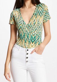 Grön och gul mönstrad kortärmad bodysuit med en djup V-ringning, parat med högmidjade vita jeans med knappdetaljer.