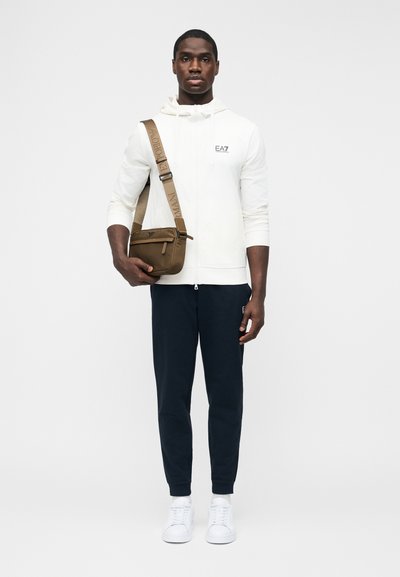 Άνδρας που φοράει λευκή φούτερ με φερμουάρ, ναυτικό μπλε φόρμα, λευκά αθλητικά παπούτσια και φέρει καφέ τσάντα χιαστί με πλατιά ιμάντα με λογότυπο.