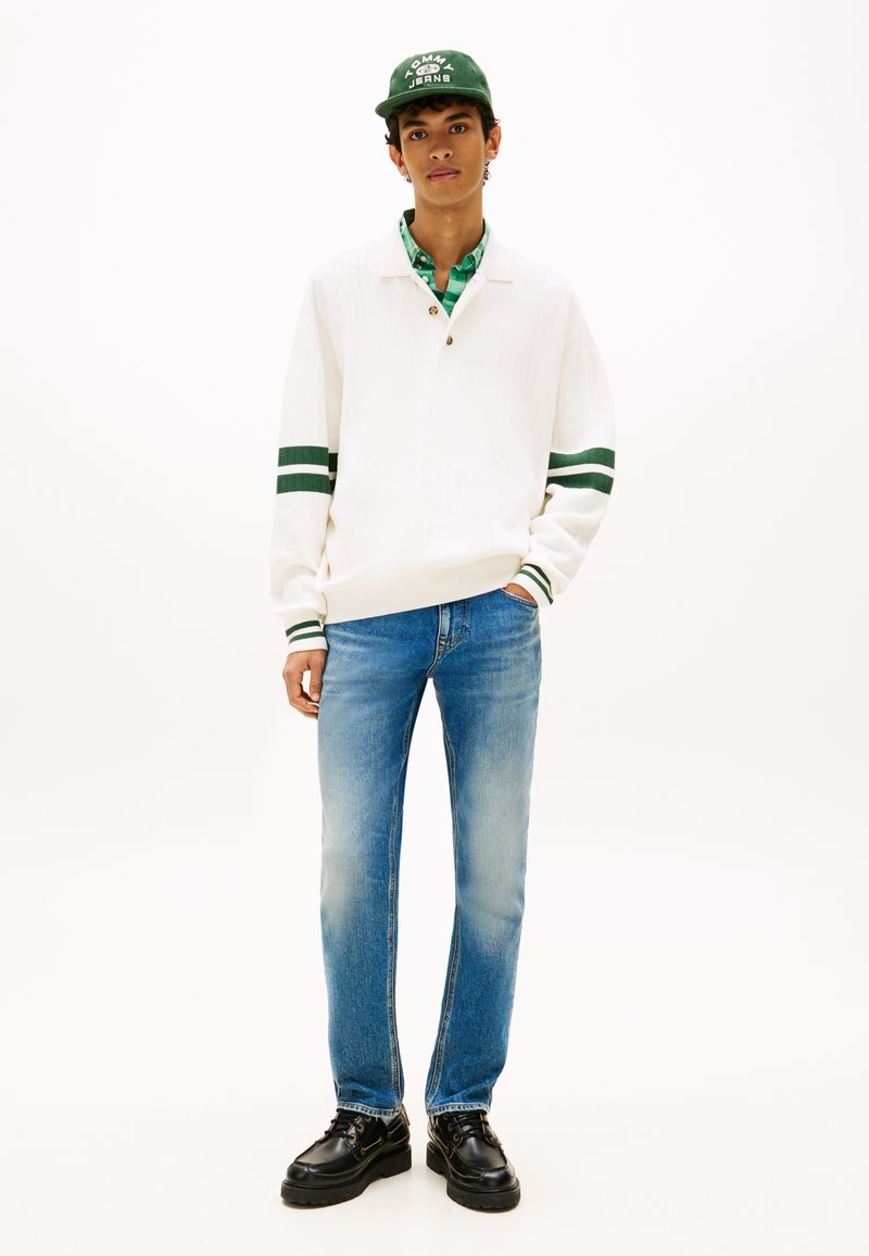 Jeune homme portant un pull blanc à rayures vertes, une chemise écossaise verte, un jean bleu, des chaussures noires et une casquette verte, debout avec une main dans la poche.