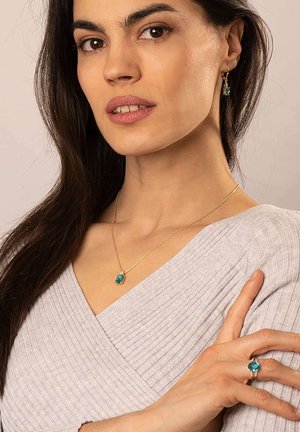 Gold necklace with a turquoise pendant, matching ring, worn by a woman in a light gray ribbed top, showcasing a smooth, elegant design.