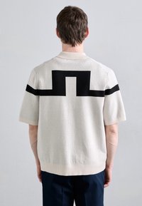 Maglietta a maniche corte di colore crema con un motivo geometrico nero sulla parte posteriore e strisce nere solide sulle spalle.