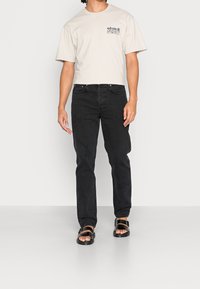 Beige Baumwoll-T-Shirt mit einem Logodesign, kombiniert mit schwarzen Jeans und schwarzen Sandalen. Die Jeans sind im geraden Schnitt mit einer klassischen Passform.