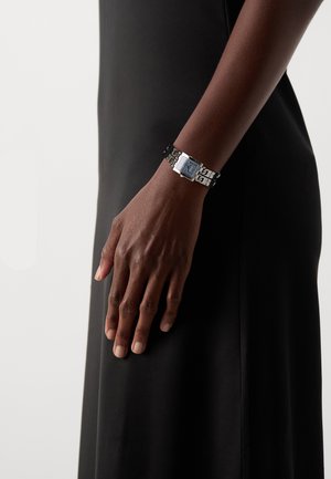 Bracelet en argent avec un cadran de montre rectangulaire, un bracelet à motifs et des détails bleus. Main reposant sur un vêtement noir uni.