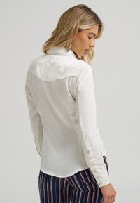 Hvit bluse med vestlig yoke med stjerne og dekorativ brodering, lange ermer med knappe detaljer, glatt stoff.