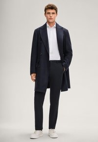 Marineblauer langer Mantel mit strukturierter Webstruktur, doppelreihigem Design und großen Revers, getragen über einem weißen Hemd und dunklen Hosen, kombiniert mit weißen Turnschuhen.