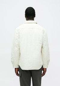 Világos bézs színű steppelt dzseki gallérral, hátul vállrész részlettel és gombos mandzsettákkal, sötétszürke nadrággal párosítva. Simított textúra, hétköznapi dizájn.