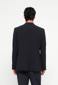 Blazer de un albastru închis, cu rever înclinat, fante unice și umeri structurați. Fabricat dintr-un material neted, cu o textură subtilă.