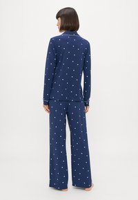 Ναυτικό σετ πιτζάμας που περιλαμβάνει μπλούζα με κουμπιά και λευκά σχέδια καρδιάς, καθώς και μακριά, χαλαρά παντελόνια με το ίδιο σχέδιο. Μαλακή υφή υφάσματος.