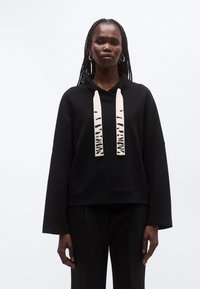 Kvinde med lange fletninger iført sort oversize sweatshirt med brede hvide snore, der har sorte abstrakte mønstre.