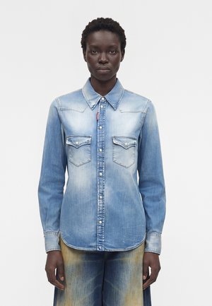 Μαύρο άτομο φοράει ένα ανοιχτό μπλε ξεθωριασμένο τζιν πουκάμισο με κουμπιά από μαργαριτάρι και τσέπες στο στήθος, στέκεται μπροστά σε ένα απλό φόντο.