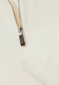 Cremfarbene Zip-up-Oberteil mit gerippter Textur, ausgestattet mit einem silbernen Metallreißverschluss und einem schwarzen Ziehband. Nahaufnahme des Kragens und des Reißverschlussbereichs.