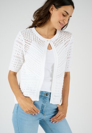 Witte gebreide cardigan met korte mouwen, met een open voorkant, een gestructureerd patroon en een centrale knoopsluiting, gecombineerd met een wit topje en denim.