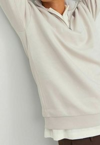 Felpa crema chiara con zip a quarto, dalla texture morbida. Presenta un colletto alto, polsini a coste e un sottogiacca bianco sotto.