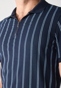 Polo shirt bleu marine avec un col zippé, arborant des rayures bleues étroites. Fabriqué en tissu doux, il possède des manches courtes et un design ajusté.