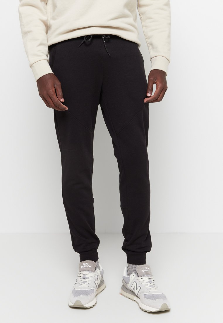 Pantalones de chándal negros hechos de una tela suave, con un corte ajustado, cinturilla con cordón y bolsillos laterales. Combinados con zapatillas grises y blancas.