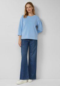 Jasnoniebieska bluza z długim rękawem i okrągłym dekoltem, zestawiona z ciemnymi, szerokimi spodniami dżinsowymi i białymi trampkami. Prosty, casualowy wygląd.