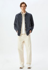 Camisa de botón azul oscuro con dos bolsillos en el pecho, llevada sobre una camisa de color claro, combinada con pantalones anchos de color blanco roto y zapatillas ligeras.