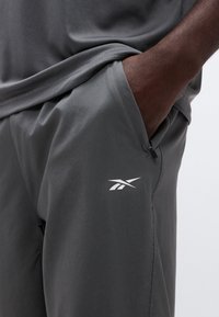 Σκούρο χέρι σε τσέπη σκούρων γκρι αθλητικών σορτς με λευκό λογότυπο Reebok, συνδυασμένο με αντίστοιχο γκρι τοπ.