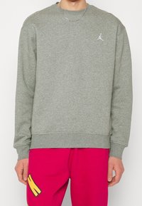 Felpa grigia con collo rotondo, polsini a coste e piccolo logo Jumpman bianco sul petto. Abbinata a pantaloni rosa con disegno giallo.