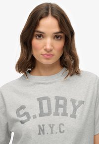 Grijze katoenen t-shirt met grote, donkergrijze tekst "S.DAY N.Y.C." in een ronde lettertype, met een ronde hals en korte mouwen.