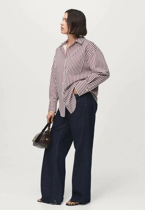 Camicia a righe rossa e bianca, abbinata a jeans in denim blu scuro a gamba larga. Tiene una piccola borsa scura e indossa sandali bassi.