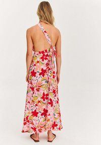 Žena oblečená v ružových kvetinových maxi šatách na jedno rameno s otvoreným chrbtom a viazaním, stojí bosá proti jednoduchému pozadiu.