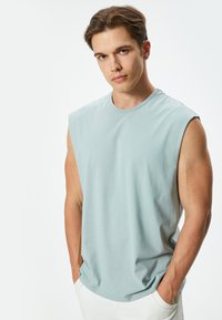 Camiseta sin mangas de color azul claro en una tela suave, con un escote redondo y un corte relajado, combinada con pantalones blancos, con las manos en los bolsillos.