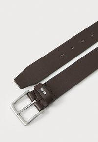 Ceinture en cuir brun avec une boucle rectangulaire en argent. Possède une texture lisse et cinq trous d'ajustement. Logo embossé près de la boucle.