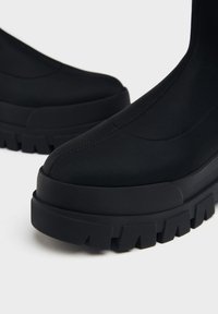 Stivali neri alla caviglia realizzati in materiale liscio ed elastico, con una suola in gomma spessa caratterizzata da profonde scanalature e un design minimalista.