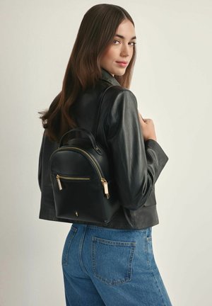 Kobieta o długich brązowych włosach, ubrana w czarną skórzaną kurtkę i niebieskie dżinsy, niesie mały czarny skórzany plecak z złotymi suwakami.